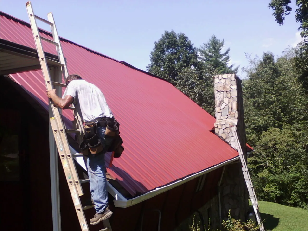 Roofer on a ladder beside a red metal roof and stone chimney — Soffit & Fascia Repair by Roofing Brothers in Wilson Creek